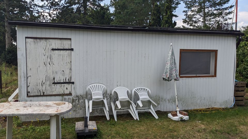 Kleines, einstöckiges Holzhaus mit flachem Dach und hellgrauer Fassade in einem Garten, umgeben von hohen Bäumen. Vor der Wand stehen gestapelte weiße Plastikstühle und ein geschlossener Sonnenschirm; links ist eine verriegelte Tür, rechts ein Fenster.