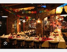 Gemütlicher Restaurantinnenraum mit Backsteinwänden und Rundbögen, langen gedeckten Tischen mit weißen Tischdecken und gefalteten Servietten. Bunte Girlanden, Ballons und Hängelampen schmücken den Raum, im Hintergrund ist ein kleiner Barbereich mit Regalen zu sehen.