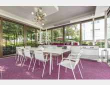 Moderner Wintergarten mit großen Glasfronten, hellen weißen Möbeln und einer offenen Küchenzeile in Weiß mit lila Akzenten. Der Boden ist mit einem lila Teppich ausgelegt, und im Hintergrund sind Bäume sowie ein Gartenbereich sichtbar.