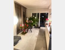 Moderner Wohnraum mit großen Bodenfliesen, bodentiefen Fenstern mit Jalousien und einer Glastür, die ins Freie führt. Im Vordergrund steht ein Adventskranz mit roten Kerzen auf einem niedrigen Ständer, daneben Pflanzen, eine Stehlampe und ein kleiner Tisch mit Getränken.