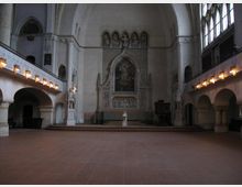 Innenansicht einer Kirche mit hohen, gewölbten Decken, großen Kirchenfenstern auf der rechten Seite und einem erhöhten Altarbereich mit Verzierungen. Der Altar ist von einem kunstvollen Rahmen mit religiösem Gemälde umgeben, flankiert von Statuen und einer Kanzel.