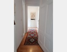 Ein schmaler Flur mit hellem Holzboden, auf dem ein langer Teppich mit buntem geometrischem Muster liegt. Am Ende des Flurs befindet sich eine weiße Tür mit einem dekorativen Kunstwerk darauf.