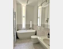 Modernes Badezimmer mit freistehender weißer Badewanne, wandhängender Toilette und Waschbecken. Wände aus hellen Fliesen, ein großer Spiegel an der rechten Seite und schmale Fenster, die natürliches Licht hereinlassen.