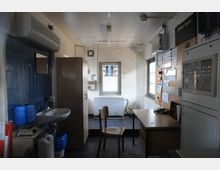 Kleiner Funktionsraum mit einem Schreibtisch, Stuhl, Waschbecken, blau gefliester Wand, Schrank und technischen Bedienelementen. An den Wänden befinden sich ein kleines Fenster, Schaltkästen und ein Klemmbrett mit aufgehefteten Papieren.