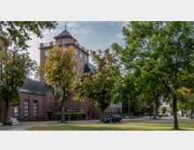Backsteingebäude mit einem hohen Turm und Spitzdach, umgeben von Bäumen, mit einem kleinen Parkplatz und einer Straße im Vordergrund. Die Szenerie zeigt eine ruhige Umgebung mit herbstlichem Laub.
