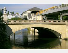 Steinbrücke über einen Fluss mit einer Statue auf der linken Seite und einer erhöhten gelben U-Bahn auf einer benachbarten Eisenbahnbrücke. Im Hintergrund sind moderne Gebäude und eine U-Bahn-Station erkennbar.