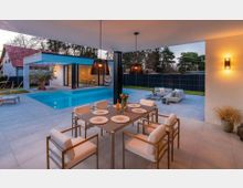 Überdachte Terrasse eines modernen Wohnhauses mit großformatigen Fliesen, Esstisch und Loungemöbeln, mit Blick auf einen rechteckigen Pool. Im Hintergrund sind bodentiefe Glasfronten, eine überdachte Sitznische, dunkler Sichtschutzzaun und Gartenbepflanzung zu sehen.