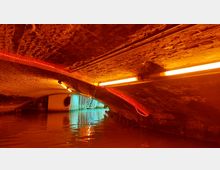 Ein gemauerter Tunnel mit niedriger Decke, beleuchtet durch orangefarbene und rote Lichter, die entlang der Wände und Decke verlaufen. Am Ende des Tunnels ist ein beleuchteter Bereich mit einer Metalltreppe sichtbar, das Wasser bedeckt den Boden des Tunnels.