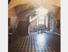 Flur in einem historischen Gebäude mit Gewölbedecke, schwarz-weiß kariertem Fliesenboden und einer hölzernen Treppe, die nach oben führt. Im Hintergrund stehen ein kleiner Tisch mit zwei Stühlen vor einem Fenster, und von der Decke hängt ein Kronleuchter.