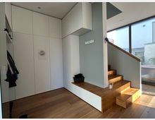 Minimalistischer Innenraum mit weißem Einbauschrank, Holzboden und moderner Holztreppe mit seitlicher Glasfassade, die natürliches Licht hereinlässt. Links ist eine Garderobe mit aufgehängten Jacken zu sehen.