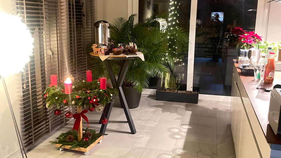 Moderner Wohnraum mit großen Bodenfliesen, bodentiefen Fenstern mit Jalousien und einer Glastür, die ins Freie führt. Im Vordergrund steht ein Adventskranz mit roten Kerzen auf einem niedrigen Ständer, daneben Pflanzen, eine Stehlampe und ein kleiner Tisch mit Getränken.