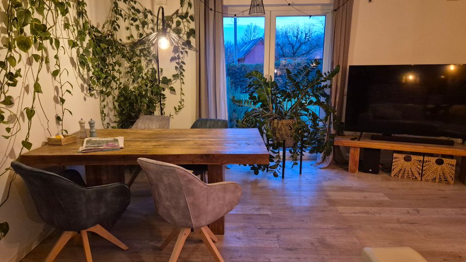 Wohnzimmer mit einem rustikalen Holztisch und vier Stühlen, umgeben von Pflanzen, die an der Wand hochranken. Im Hintergrund bodentiefe Fenster mit Blick auf einen Garten, daneben ein Fernseher auf einem niedrigen Holztisch mit Körben darunter.