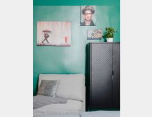 Wohnzimmer mit grün gestrichener Wand, dekoriert mit mehreren bunten Gemälden. Im Vordergrund steht ein helles, gepolstertes Sofa mit Kissen und Decke, daneben ein schwarzer Schrank mit perforierten Türen und eine kleine Topfpflanze darauf.