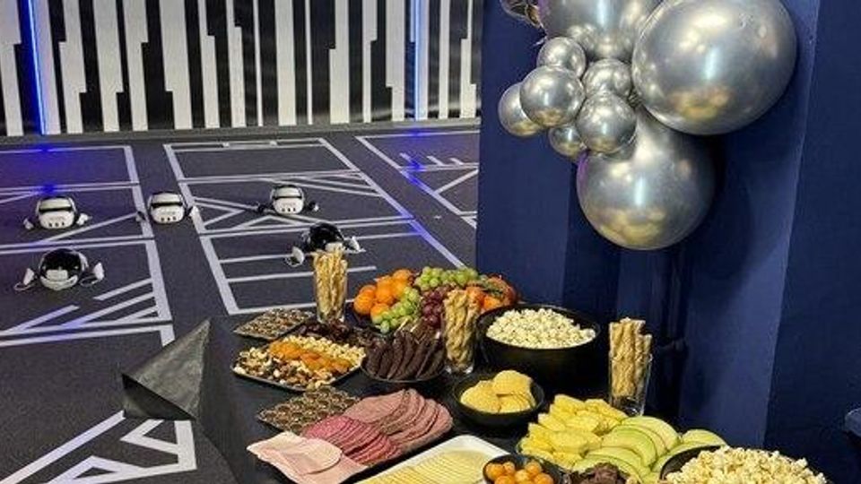Buffet-Tisch mit Käse-, Wurst- und Obstplatten sowie Snacks vor einer dunkelblauen Wand mit silberner Ballondekoration; im Hintergrund ein Indoor-Spielbereich mit markierten Spielfeldern und kleinen Roboterfahrzeugen auf dem Boden.