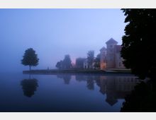 Ein Schlossgebäude mit Türmen am Ufer eines ruhigen Sees, umgeben von Bäumen und in dichten Nebel gehüllt, der die Umgebung weichzeichnet. Die Szene zeigt spiegelnde Reflexionen der Landschaft im Wasser und ein gedämpftes Licht, das aus einem Eingang scheint.