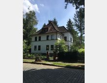 Zweigeschossiges Wohnhaus mit weißer Fassade und dunkelbraunem Walmdach, umgeben von Bäumen und einem Garten mit Zaun und Tor aus Metall und Ziegelpfeilern. Im Vordergrund ist eine gepflasterte Straße zu sehen.