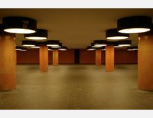 Unterirdischer Durchgang mit orange gefliesten Rundsäulen, dunkelgrauem Boden und an der Decke montierten schwarzen Leuchten mit kreisförmiger, heller Beleuchtung. Die Wände sind mit orangefarbenen Fliesen verkleidet.