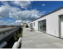 Moderne Dachterrasse mit großen grauen Fliesen, angrenzend an ein weißes Gebäude mit Glas-Schiebetüren. Im Hintergrund sind Begrünungselemente, ein Tisch mit Stühlen und umliegende Dächer unter einem teils bewölkten blauen Himmel zu sehen.
