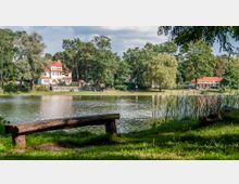 Ein kleiner See mit grasbedecktem Ufer, im Vordergrund eine einfache Holzbank und im Hintergrund eine Villa mit rotem Dach sowie ein weiteres Gebäude neben Bäumen. Die Szenerie ist von dichtem Grün und lockerer Bebauung umgeben.
