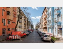 Straßenansicht einer Wohngegend mit mehrstöckigen Häusern in verschiedenen Farben wie Orange, Gelb und Blau. Gepflasterte Straße mit parkenden Autos auf beiden Seiten, vereinzelt Bäume und ein blauer Himmel mit weißen Wolken im Hintergrund.