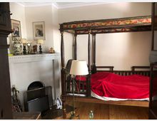 Zimmer mit einem hölzernen Himmelbett, das eine rot gemusterte Verzierung am Baldachin und eine rote Decke hat. Im Hintergrund ein weißer Kamin mit dekorativen Elementen, darunter Lampen und Vasen, sowie ein Holzboden und verschiedene Gegenstände wie ein Standspiegel und ein Bügeleisen.