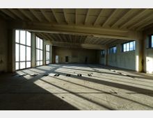 Großer, leerer Raum mit Betonboden, hohen Decken und einer statischen Struktur aus sichtbaren Trägern. Große Fenster auf der linken und rechten Seite lassen Tageslicht einfallen, während der Raum an einigen Stellen beschädigte oder fehlende Bodenplatten aufweist.