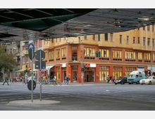 Straßenszene mit einem großen orangefarbenen Eckgebäude, darunter Geschäfte mit Schaufenstern und einem Bekleidungsgeschäft. Im Vordergrund befinden sich Verkehrsschilder, eine Ampel, Autos, Fahrräder und Fußgänger, teils unter einer Brücke.