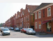 Reihe historischer Backsteinhäuser mit Treppengiebeln entlang einer gepflasterten Straße, auf der Autos und Fahrräder geparkt sind; einige Häuser haben Schilder für Geschäfte oder Praxen.