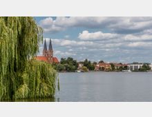 Blick auf eine Seeuferlandschaft mit einer großen Trauerweide im Vordergrund. Im Hintergrund sind eine Kirche mit zwei spitzen Türmen, Wohnhäuser und Bäume zu sehen, unter einem bewölkten Himmel.