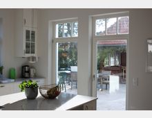 Moderne Küche mit weißem Interieur und großen Fenstertüren, die in einen sonnigen Außenbereich mit Terrassenmöbeln und grüner Vegetation führen. Auf einer Kücheninsel stehen eine Pflanze in einem Topf und eine Obstschale.