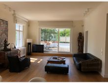 Wohnzimmer mit Holzboden und großen Fenstern, die zu einer Terrasse mit Gartenmöbeln führen. Der Raum ist mit schwarzen Ledersesseln, einem niedrigen Holztisch, Kunstobjekten und einer stehenden Holzskulptur dekoriert.