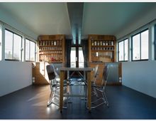 Innenraum eines schmalen Raumes mit großen Fenstern an den Seiten, eingerichtet als minimalistische Küche mit holzverkleideten Schränken. An den Rückwänden befinden sich Regale mit Gläsern, Messern und anderen Küchengeräten, in der Mitte steht ein Tisch mit Metallstühlen.