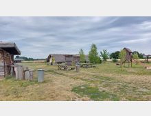 Weitläufiger Außenbereich mit Holzhütten und Strohdächern, rustikalen Holzbänken und Tischen sowie einem kleinen Spielhaus mit Rutsche auf einer Wiese. Im Hintergrund sind Bäume und offene Felder zu sehen.