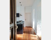 Blick in eine helle Küche mit Holzboden und weißen Wänden. Zu sehen sind ein schwarzes Küchenelement mit Obstschale, eine weiße Tür, Fliesen im U-Bahn-Stil, hängende Lampen sowie ein Durchgang mit weiß gestrichenen Backsteinbögen.