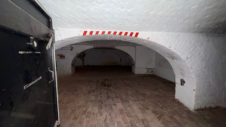 Gewölbekeller mit weiß gestrichenen Ziegeldecken und Ziegelboden, gesehen durch eine geöffnete schwarze Metalltür. Im Hintergrund führt ein weiterer niedriger Rundbogen in einen dunkleren Nebenraum; an der Decke sind rot-weiße Markierungen angebracht.