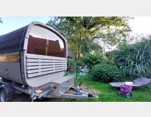 Ein mobiler Sauna-Anhänger mit gebogenem Dach und Holzverkleidung steht auf einer Wiese in einem Garten mit hohen Bäumen und dichter Vegetation. Im Vordergrund befinden sich Gartenmöbel, eine Sonnenliege und ein Kinderwagen.