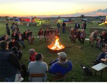 Eine Gruppe von Menschen sitzt in einem Kreis um ein offenes Lagerfeuer auf einer Wiese. Im Hintergrund sind mehrere kleine bunte Flugzeuge und ein Sonnenuntergang am Horizont zu sehen.