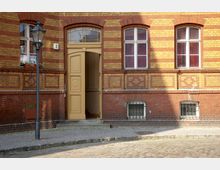 Backsteinfassade eines historischen Gebäudes mit gelben und roten Ziegeln, großen rechteckigen Fenstern mit weißen Rahmen und einem offenen, hellen Eingangstor. Eine Laterne steht am Gehweg vor dem Gebäude, und der Boden besteht aus Kopfsteinpflaster.