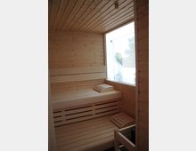 Holzverkleideter Sauna-Innenraum mit Sitz- und Liegeflächen aus hellem Holz. Rechts befindet sich ein großes Fenster mit Blick ins Freie, das Tageslicht in den Raum einlässt.