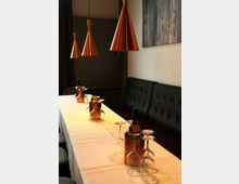 Langer, schmaler Restauranttisch mit weißer Tischdecke, gedeckt mit umgedrehten Weingläsern und Flaschen in Metallkühlern. Darüber hängen mehrere kupferfarbene Pendelleuchten, daneben verläuft eine dunkel gepolsterte Sitzbank entlang der Wand mit einem Wandbild.