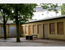 Ein gepflasterter Innenhof mit Bäumen, umgeben von einem beige-gelben Gebäude mit heruntergelassenen Rollläden und vereinzelten Graffiti. Vor dem Gebäude stehen eine Holzbank und ein kleiner Tisch.