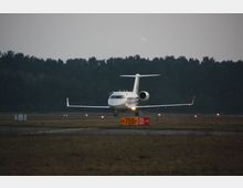 Ein kleines Passagierflugzeug steht auf einer Landebahn eines Flughafens, umgeben von einem Wald im Hintergrund. Im Vordergrund ist ein gelb-rotes Schild mit den Markierungen „750 m“ und „05“ zu sehen.