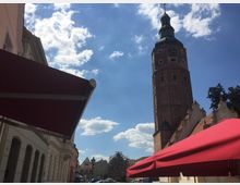 Straßenszene mit Blick auf einen roten Backsteinturm mit einer Uhr und grüner Kuppel, umgeben von historischen Gebäuden. Im Vordergrund befinden sich rote Markisen und ein klarer Himmel mit wenigen Wolken.