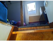 Ein kleiner Dachboden mit einer Holztreppe, der teilweise als Abstellraum genutzt wird, sichtbar durch Kartons, Gegenstände und eine Lagerbox. Ein schräges Fenster lässt Tageslicht herein, und ein Holzpfosten steht in der Mitte des Raums.