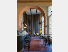 Ein modernes Café oder eine Bar mit hohen Decken, sichtbaren Rundbögen und großen Fenstern, die viel Licht hereinlassen. Links im Bild befindet sich eine lange Theke mit Hockern, rechts stehen kleine Tische und Stühle, während im Hintergrund lange Vorhänge und Pflanzen den Raum dekorieren.