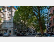 Straßenszene mit mehrstöckigen Wohngebäuden im Altbaustil, Bäumen, die die Straße säumen, und parkenden Autos entlang der Bordsteine. Im Erdgeschoss eines Gebäudes befinden sich Geschäfte mit Schaufenstern.