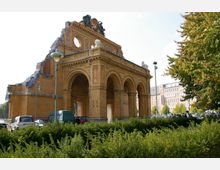 Ziegelsteinruine eines historischen Gebäudes mit einem zentralen Rundfenster, drei großen Rundbögen und dekorativen Skulpturen auf dem Dach. Im Vordergrund sind Büsche, parkende Autos und Laternen, im Hintergrund moderne Gebäude und Bäume.