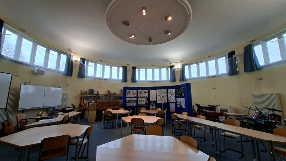 Runder Klassen- oder Musikraum mit umlaufender Fensterfront im oberen Wandbereich und einer kreisförmigen Deckenleuchte. Im Raum stehen mehrere Gruppentische und Stühle, an den Wänden befinden sich Whiteboards, Regale und Instrumente wie Klavier, Keyboard und Schlagzeug.