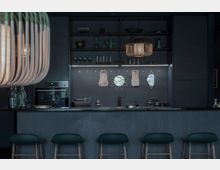 Moderne Küche mit dunklem Design, schwarzer Küchenzeile und einer Bar mit vier Hockern. Die Rückwand ist mit Regalen, kleinen Dekorationsgegenständen sowie geometrischen Spiegeln verziert, und zwei hängende Holzlampen beleuchten den Raum.
