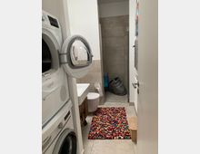 Kleines Badezimmer mit Waschmaschine und Trockner an der linken Wand, einem Waschbecken und WC in der Mitte sowie einer gefliesten Dusche im Hintergrund. Ein bunter Teppich liegt vor dem Waschbecken, und ein Holzhocker steht rechts neben einer Heizung.